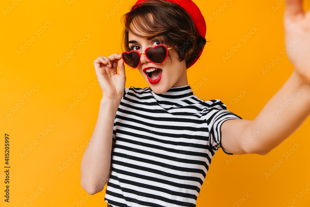 Glamorous french woman posing in red beret. Studio shot of brunette girl in striped t-shirt isolated on yellow background. Glamorous french woman posing in red beret. Studio shot of brunette girl in striped t-shirt isolated on yellow background.