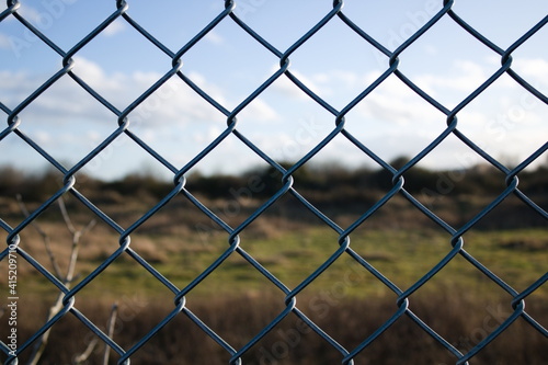 Chain link wire fence close up with blurred foliage in the background, winter, blue sky, UK