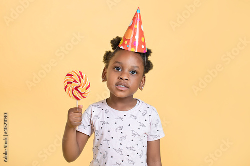 Close up of little adorable african american girl in party hat holding lollipop sweet in hands, posing to camera over yellow background, Copy Space
