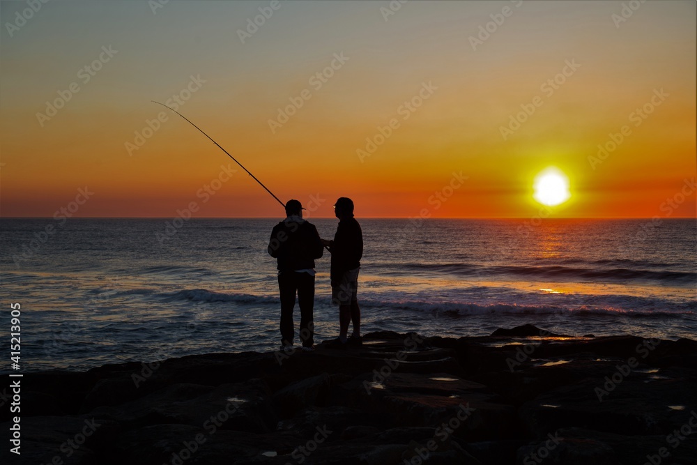 Silueta de dos pescadores conversando durante una puesta de sol con colores amarillos y naranjas sobre el Océano Atlántico en Esmoriz (Portugal)