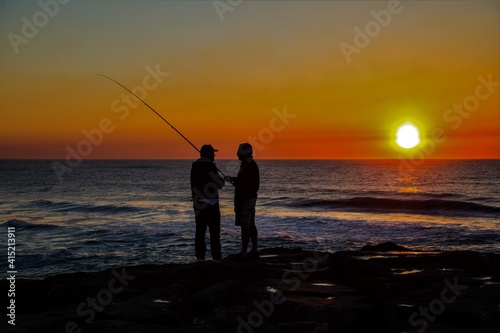 Silueta de dos pescadores conversando durante una puesta de sol con colores amarillos y naranjas sobre el Océano Atlántico en Esmoriz (Portugal)