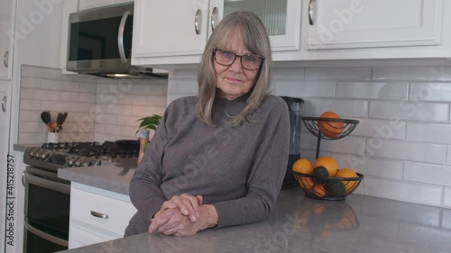 Medium shot portrait of grey haired senior woman with glasses looking at camera in her kitchen. Older Caucasian retiree in a gray sweater. Slow motion 4k
