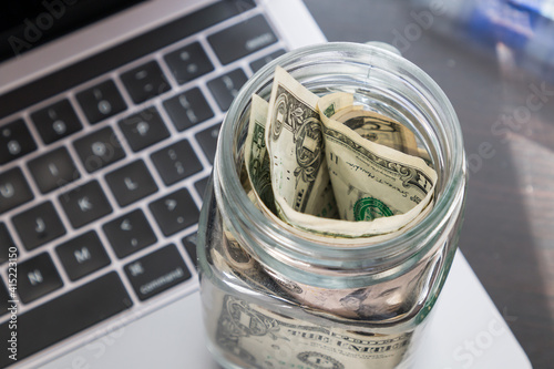 Overhead shot of a glass jar filled with bills including USD cash bills and coins in 5 and 1 dollar denominations on a modern laptop computer keyboard to display finances and savings