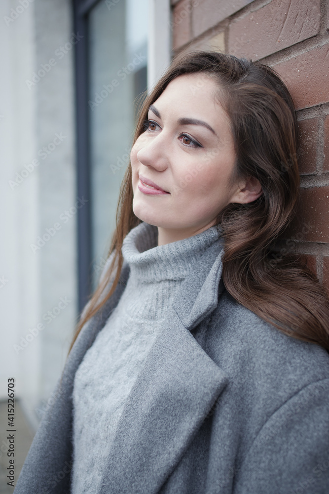 Pin-up girl posing on the street for a walk. A beautiful girl with long dark hair, a gray long coat and a cozy warm gray sweater is leaning against a brick wall in the street and smiling mysteriously