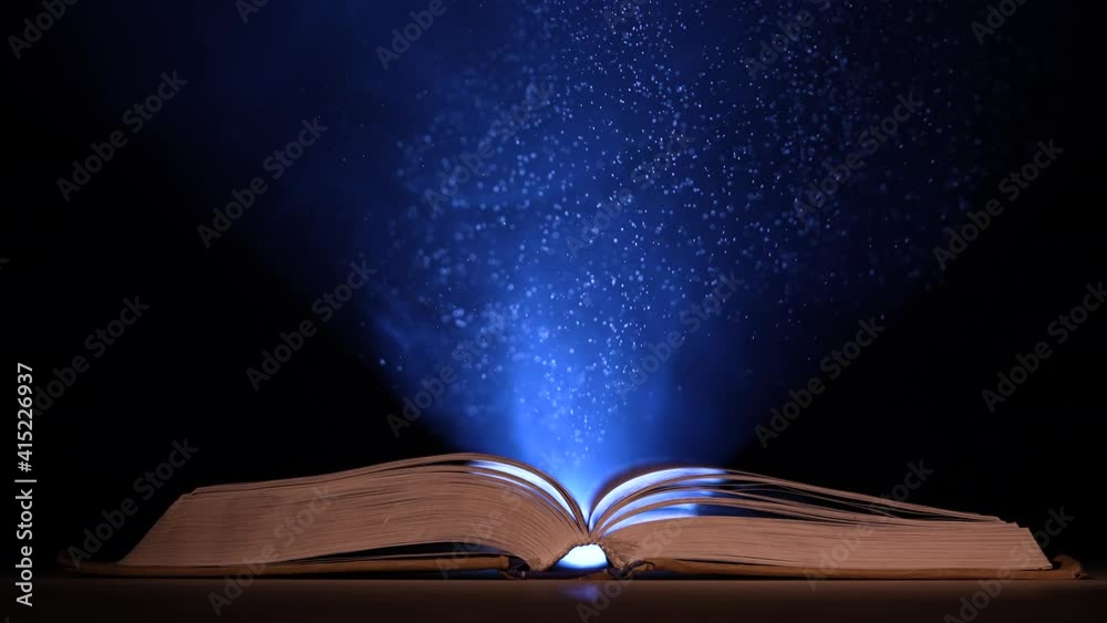 In a dark, smoky studio, a thick, open book lies on a table against a black background. Particles of dust in the light of a searchlight fly up over an open book. Slow motion. Close up.