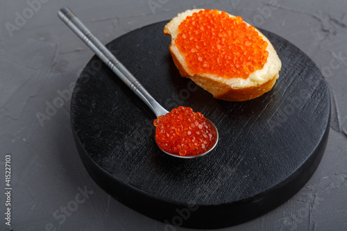 One bruschetta with butter and red caviar next to a spoon with caviar on a wooden board on a concrete background.