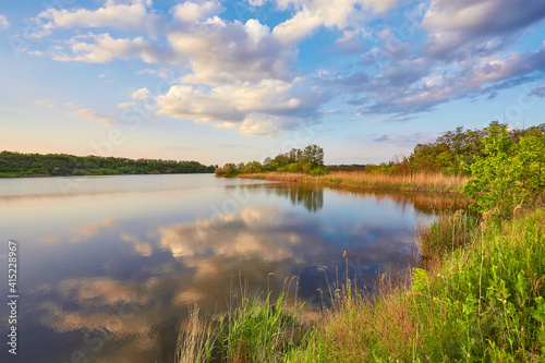 Fototapeta Naklejka Na Ścianę i Meble -  Beautiful river coast at sunset in summer. Colorful landscape with lake, green trees and grass, blue sky with multicolored clouds and orange sunlight reflected in water.