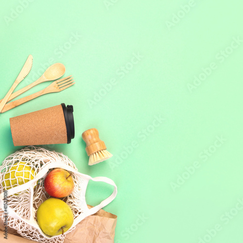 Reusable wooden cutlery, cork mug and grocery bag with apples. Dishwashing brush and kraft wrapping paper, eco friendly fork, knife, spoon on green mint square backdrop. Zero waste concept. Copyspace.