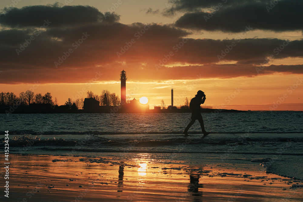Silhouette of unrecognizable person walking on Mangalsala pier through the waves in sunset. Orange sky and silhouettes of lighthouse in the background
