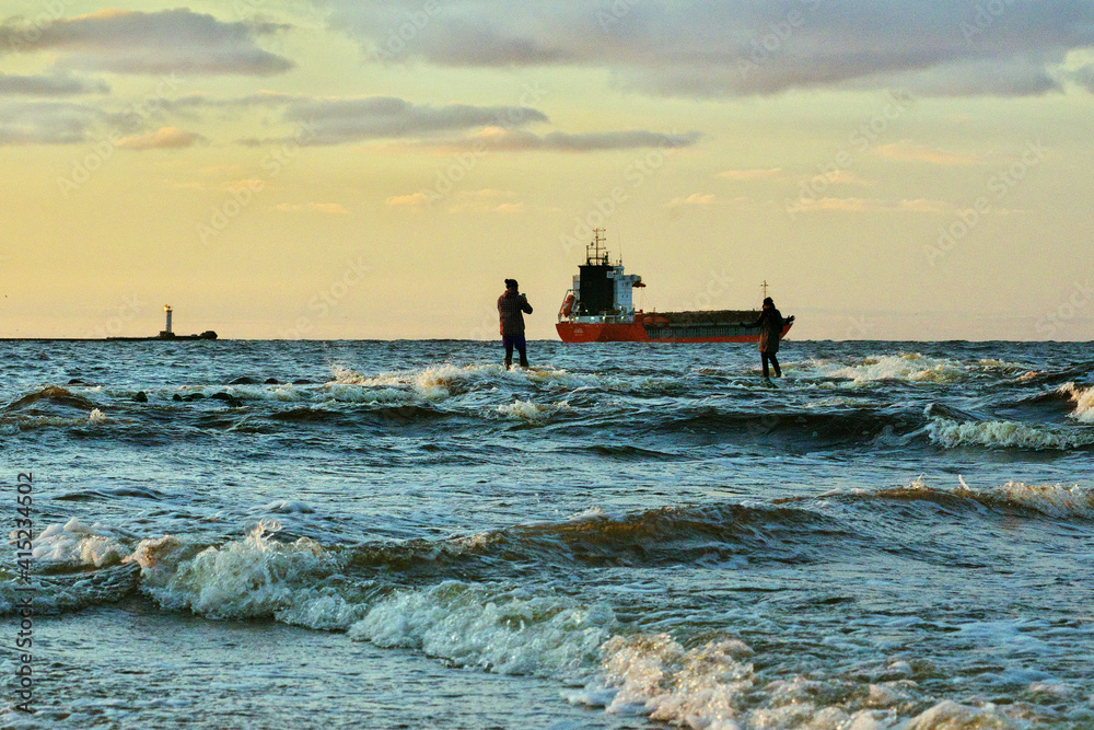 
Two unrecognizable persons taking photo on Mangalsala pier in sunset. Yellow and blue sky, Mangalsala lighthouse and red ship in the background