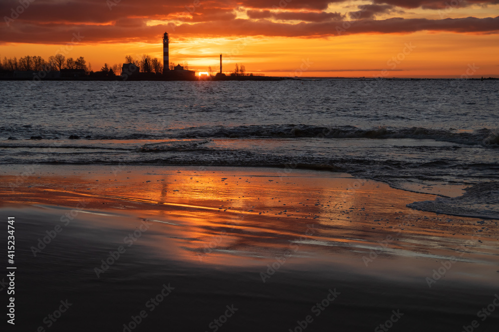 Beautiful feflections of orange sunset sky in seaside water in Mangalsala. Daugavgriva lighthouse with sun on the horizon in background. Focus on foreground