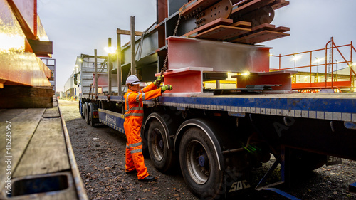 Driver Wearing Full PPE and Chaining Down a Load of Steel Structures onto a Semi Truck Flat bed with Metal Posts Fitted.