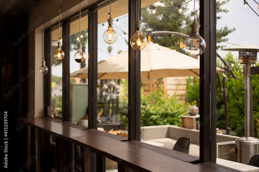 A view from inside a cafe setting, looking through a wall of windows towards a comfortable isolated patio area, featuring vintage hanging light bulbs.