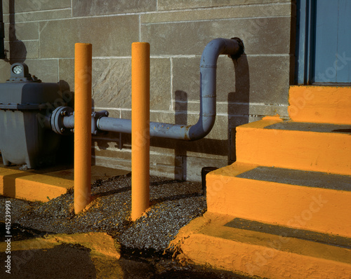 A minimalistic urban industrial geometric study of Bright yellow steps, protection poles, and a  gray gas meter at the rear door of the post office in Bllomington, Indiana 1986.