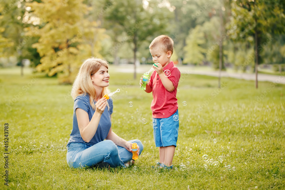 Young Caucasian mother and boy toddler son blowing soap bubbles in park. Mom and child playing having fun together outdoor on summer day. Happy authentic family childhood lifestyle.