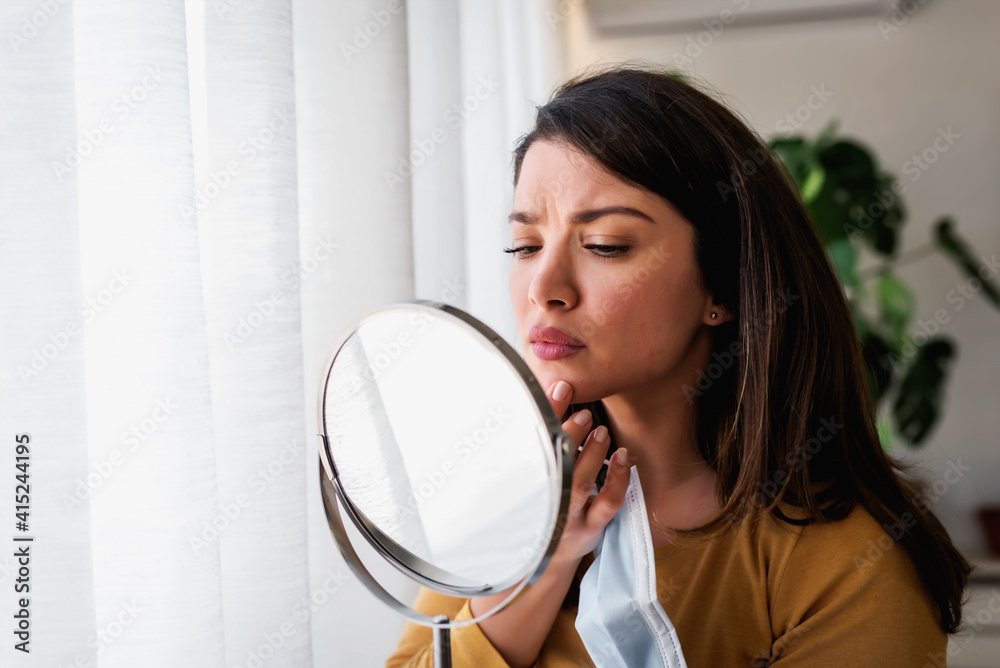 Woman looking herself in the mirror at home standing by the big window. She is concerned about acne, maskne Woman looking herself in the mirror at home standing by the big window. She is concerned about acne, maskne
