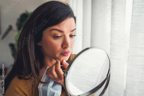 Woman looking herself in the mirror at home standing by the big window. She is concerned about acne, maskne photo