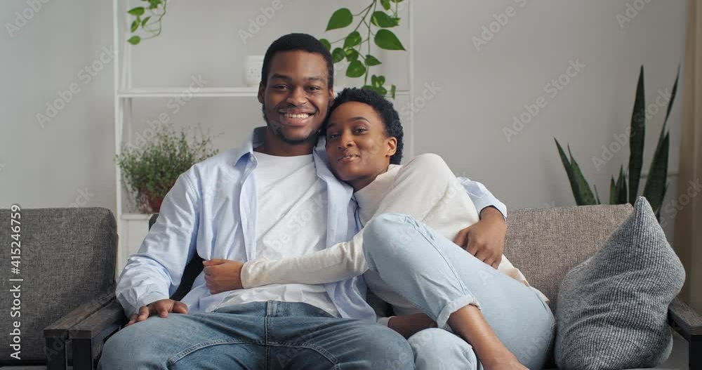Afro american couple wears casual clothes sitting together at home living room interior happy newlyweds hugging on couch smiling looking at camera spending time together, weekend relax, love concept Afro american couple wears casual clothes sitting together at home living room interior happy newlyweds hugging on couch smiling looking at camera spending time together, weekend relax, love concept