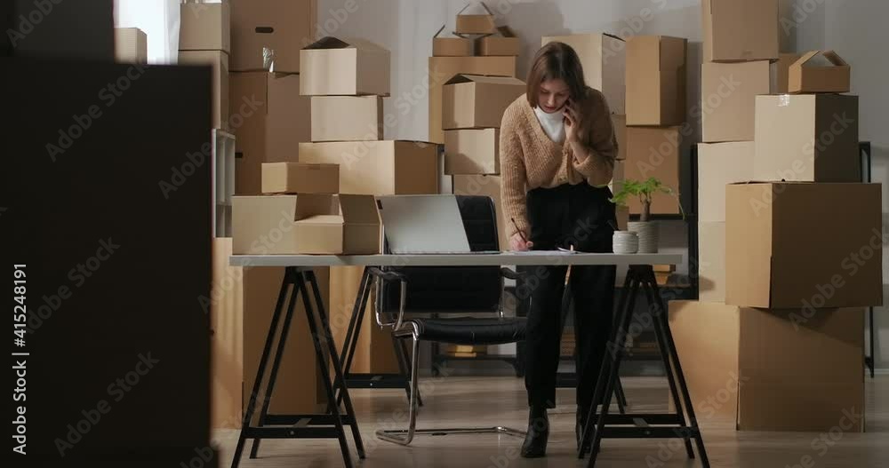 woman owner of small delivery company is communicating by phone with supplier in room of flat using as warehouse with boxes
