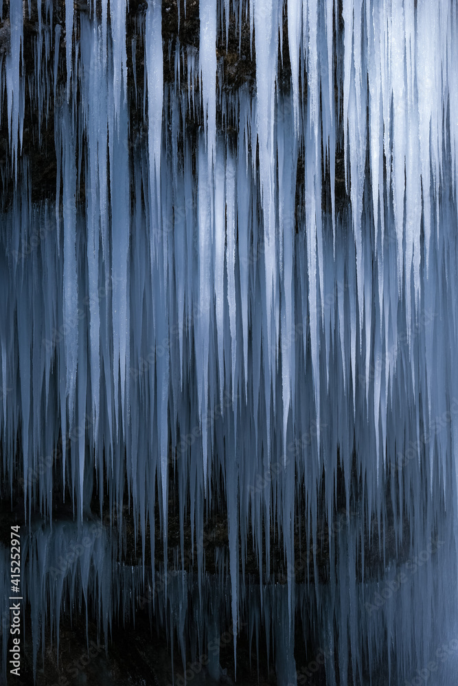 Beautiful long icicles of a frozen waterfall, with water flowing and crashing down and Ice water dripping from the tips of icicles in a cold eery and moody atmosphere in a cave in the mountains 
