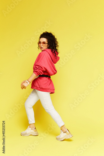 A young woman in sportswear and sunglasses over a yellow background. Young beautiful woman in a pink hoodie and white pants at the yellow background, isolated