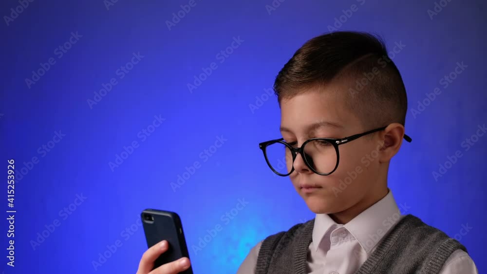 Handsome boy with glasses looks is surprised at smart phone at blue background. Modern child is joy looking at gadget. Handsome boy with glasses looks is surprised at smart phone at blue background. Modern child is joy looking at gadget.
