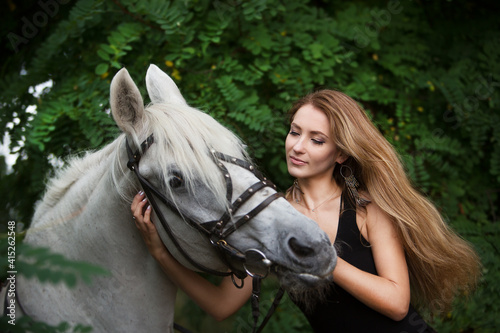 Young beautiful woman with long hair hugs her horse while walking in the forest.