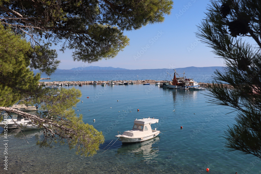 Small pier with boats on the Croatian sea coast on a sunny day against the backdrop of blue sky and green pine trees