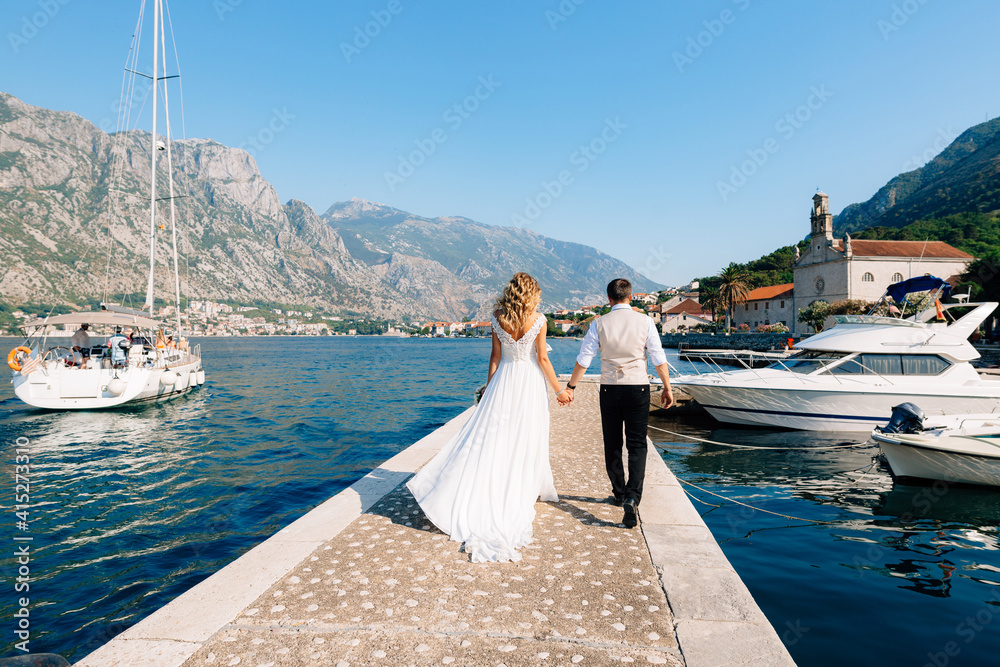 The bride and groom are walking along the pier in the Bay of Kotor, next to them are boats, yachts and sailing boat