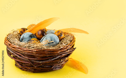 Easter colored eggs in a basket in the form of a nest on a yellow background. Decorated with orange feathers. Close up, copy space. Easter holiday concept