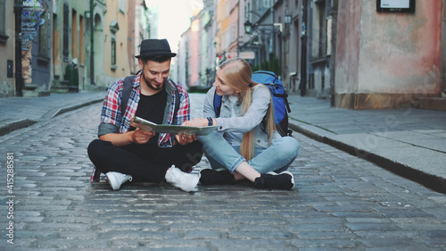 Young tourists couple using map, sitting on pavement and admiring historical surroundings. They are excited and smiled. © art24pro