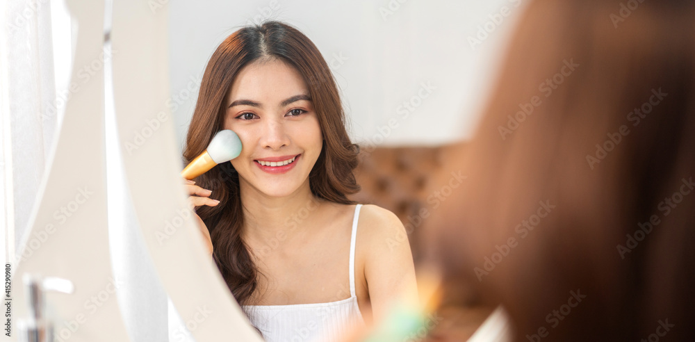 Smiling of young beautiful pretty asian woman clean fresh healthy white skin looking at mirror.asian girl holding make-up brushes and make up on face with cosmetics set at home.facial beauty Smiling of young beautiful pretty asian woman clean fresh healthy white skin looking at mirror.asian girl holding make-up brushes and make up on face with cosmetics set at home.facial beauty