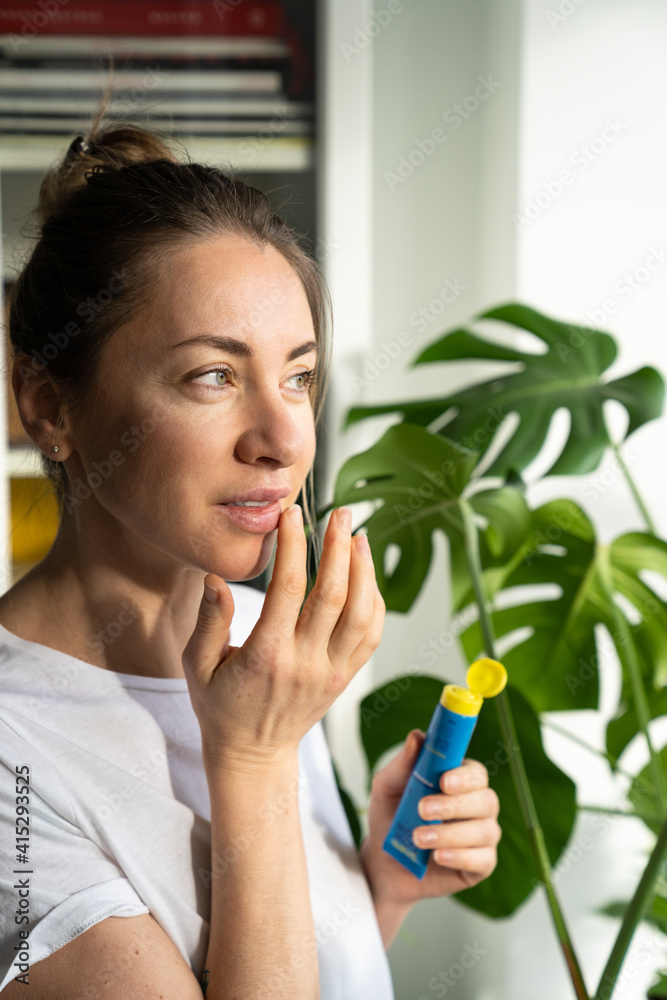 Woman applying moisturizing nourishing balm to her lips with her finger to prevent dryness and chapping in the cold season, standing and looking at window. Lip protection. Woman applying moisturizing nourishing balm to her lips with her finger to prevent dryness and chapping in the cold season, standing and looking at window. Lip protection.