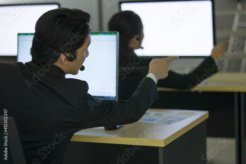 Asian man and woman officer wearing headphone headset and working with happy face in call center in front of computer screen in office at night. Overtime, late work and 24 hours service concept