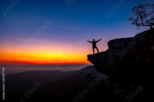 Silhouette of man standing a lone on top cliff of mountain with orange sunrise in the morning light from the back and show two hands.Concept to create energy and positive thinking.