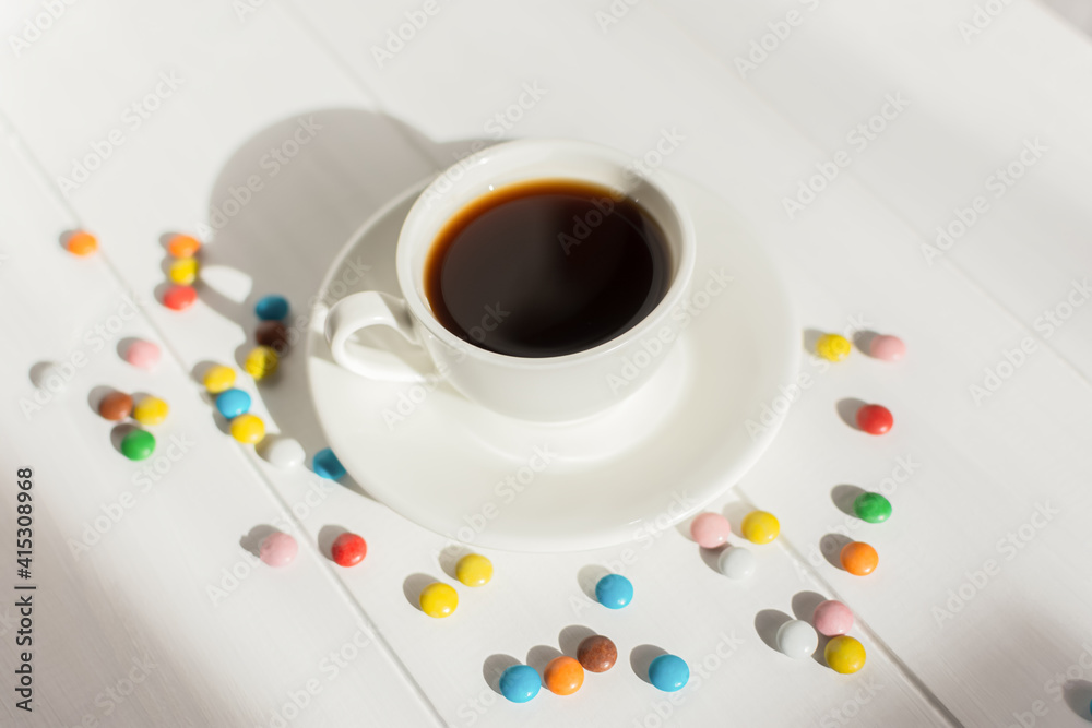 Espresso in a white cup on the table in bright sunlight. Creative composition with colorful candies. Modern breakfast. The concept of freshness of a summer morning.Minimalism, top view, white on white Espresso in a white cup on the table in bright sunlight. Creative composition with colorful candies. Modern breakfast. The concept of freshness of a summer morning.Minimalism, top view, white on white