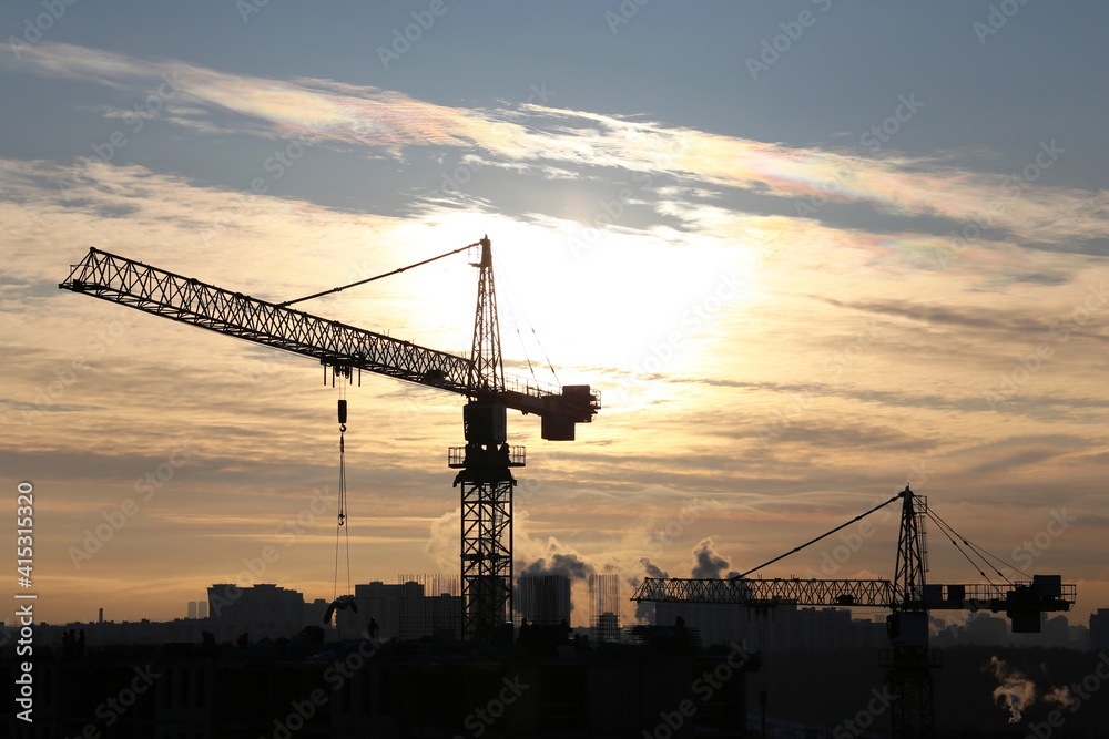 Silhouettes of construction cranes and residential buildings on sun background. Housing construction, apartment block in winter city