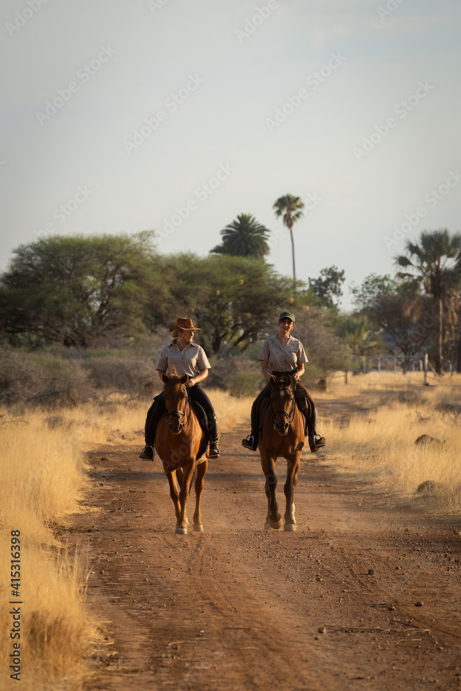 Naklejka premium Two horsewomen ride side-by-side along dirt track