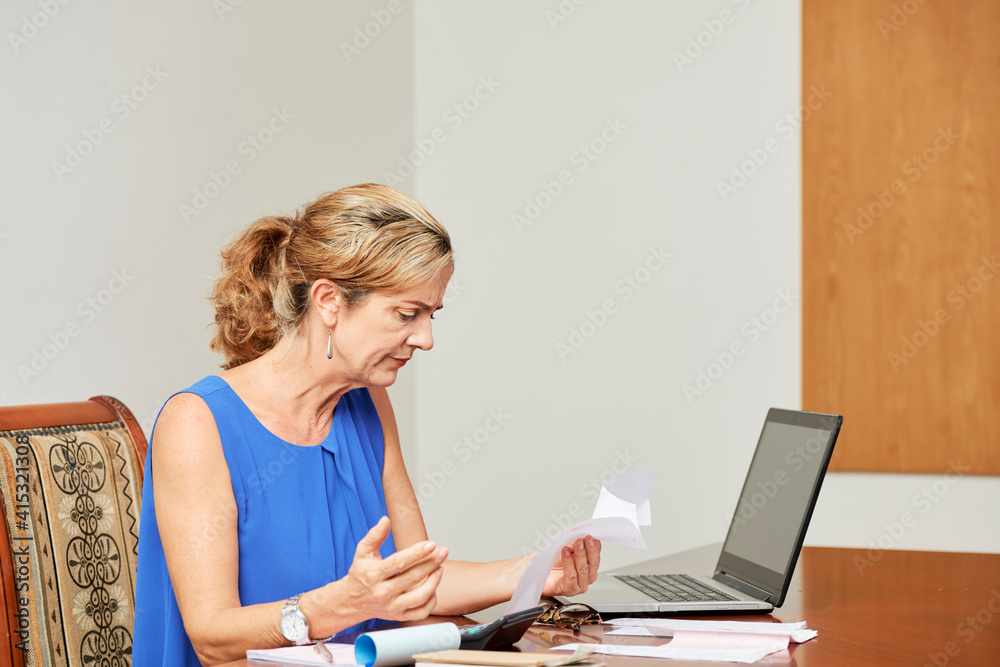 Frowning mature woman checking bills and checks when managing household finances Frowning mature woman checking bills and checks when managing household finances