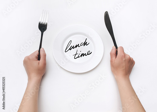 Female hands hold a fork and knife near a white plate on which the words - Lunch time is written. The concept of a balanced diet, ration and medical fasting. Top view, white background, copy space.