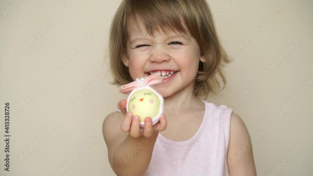 Little girl holds chicken egg decorated for chick Easter with bow. Happy child with handmade painted egg looks at camera and smiles. Easter celebration. Little girl holds chicken egg decorated for chick Easter with bow. Happy child with handmade painted egg looks at camera and smiles. Easter celebration.