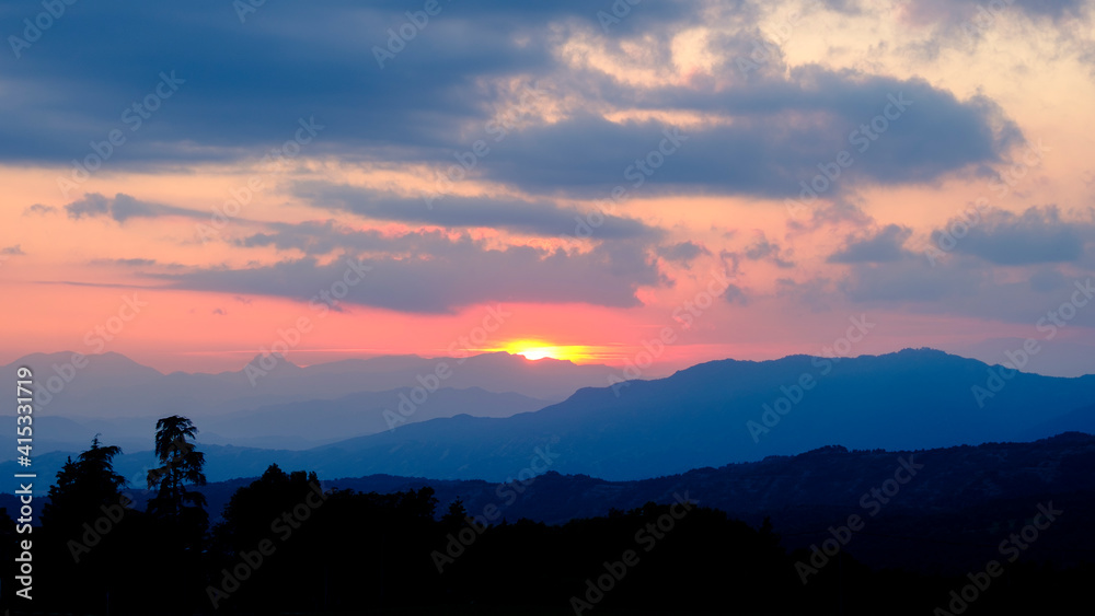 View of trees on a hill at cloudy sunset with fog light in the misty mountains at the background.