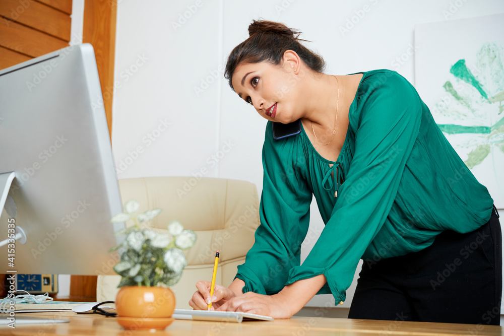 Pretty female entrepreneur leaning on office table, talking on phone with coworker or customer and writing in planner Pretty female entrepreneur leaning on office table, talking on phone with coworker or customer and writing in planner