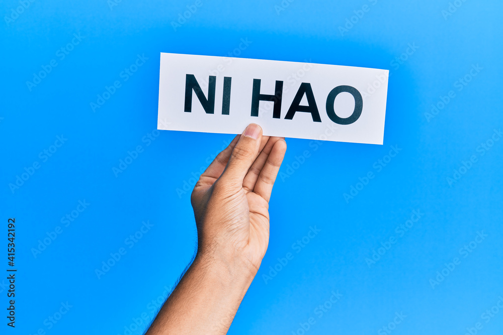 Hand of hispanic man holding chinese greeting ni hao word paper over isolated blue background. Hand of hispanic man holding chinese greeting ni hao word paper over isolated blue background.