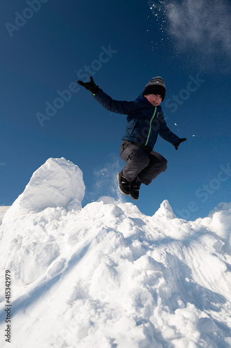 Little cheerful boy in sportswear jumps from a white snowy mountain against a blue sky.