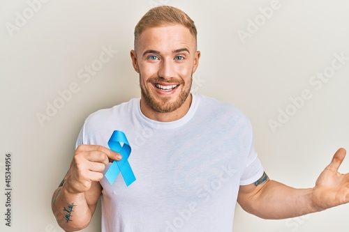 Young caucasian man holding blue ribbon celebrating achievement with happy smile and winner expression with raised hand