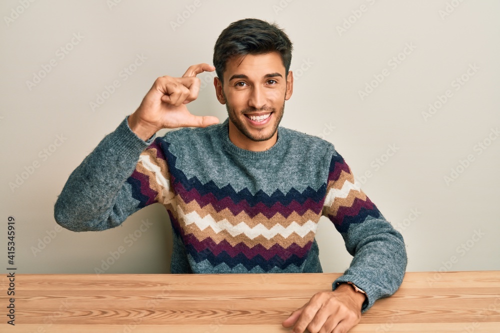 Young handsome man wearing casual sweater sitting on the table smiling and confident gesturing with hand doing small size sign with fingers looking and the camera. measure concept. Young handsome man wearing casual sweater sitting on the table smiling and confident gesturing with hand doing small size sign with fingers looking and the camera. measure concept.
