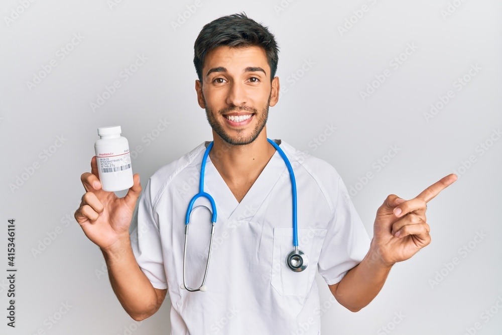 Young handsome man wearing doctor uniform holding presciption pills smiling happy pointing with hand and finger to the side Young handsome man wearing doctor uniform holding presciption pills smiling happy pointing with hand and finger to the side