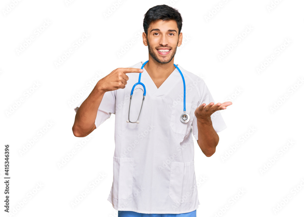 Young handsome man wearing doctor uniform and stethoscope amazed and smiling to the camera while presenting with hand and pointing with finger. Young handsome man wearing doctor uniform and stethoscope amazed and smiling to the camera while presenting with hand and pointing with finger.