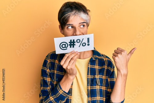Young hispanic man covering mouth with insult message paper pointing thumb up to the side smiling happy with open mouth photo